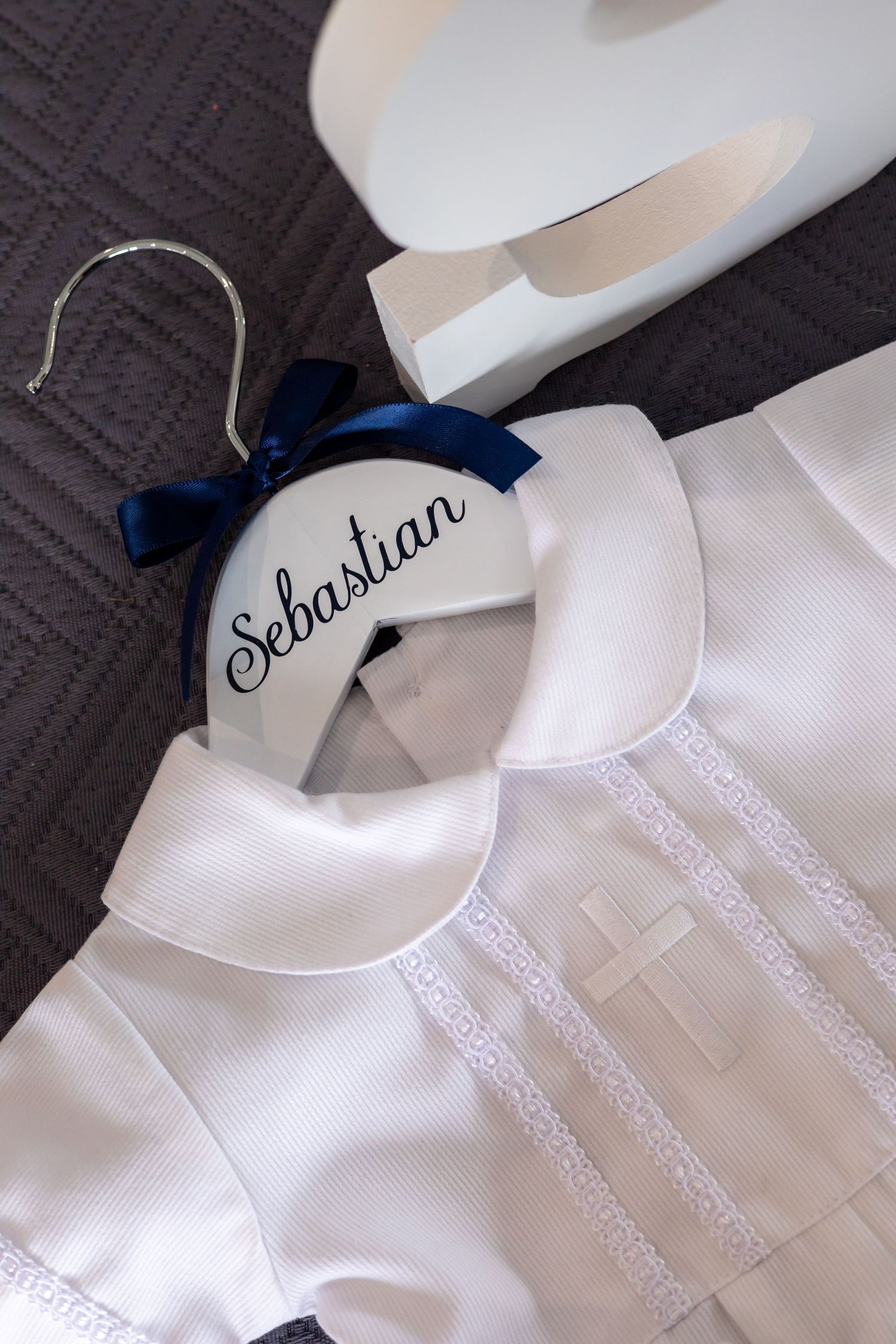 White christening gown with embroidered cross and personalized hanger with navy blue bow displaying 'Sebastian' name.