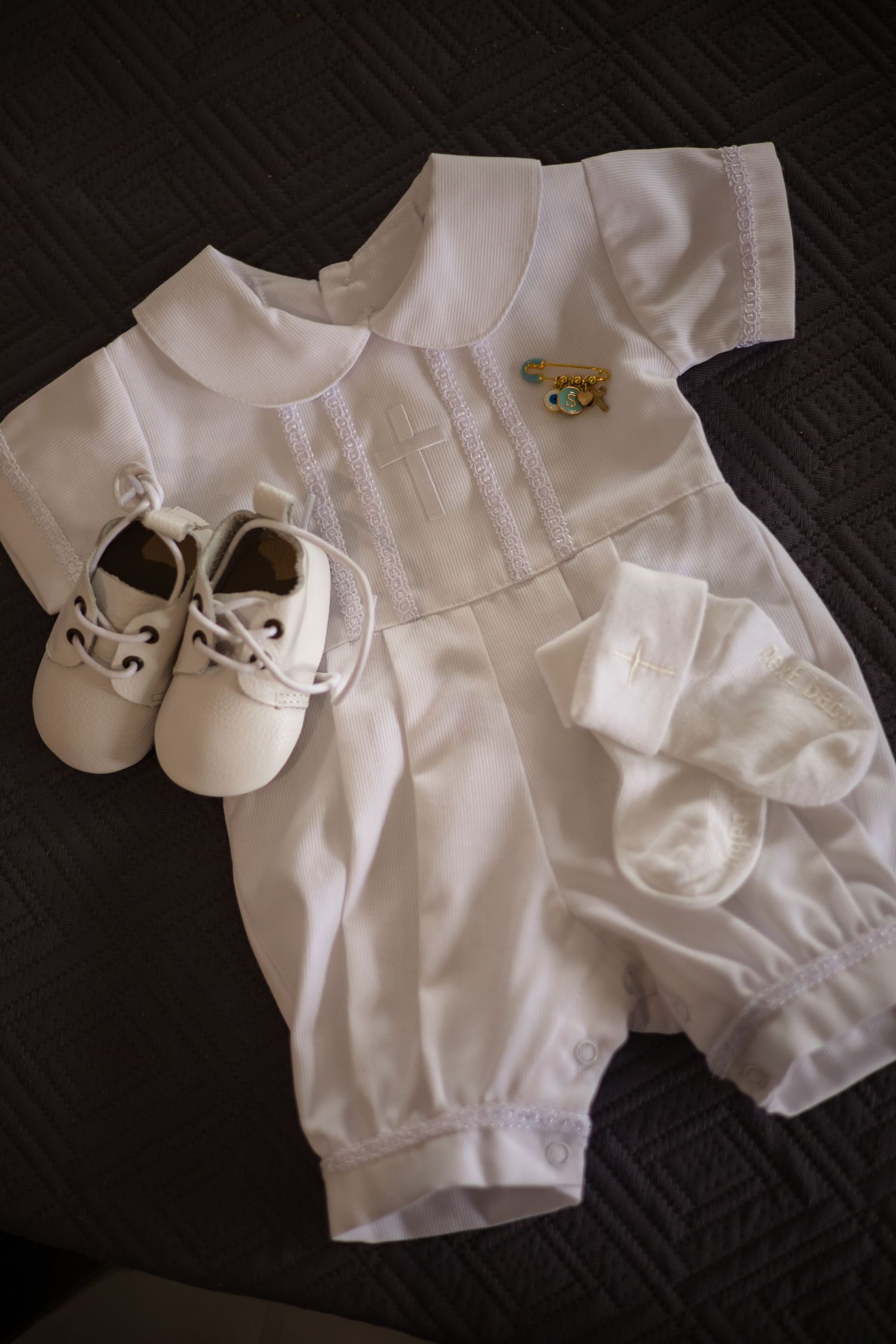 Vintage white baby christening romper with socks and tiny leather shoes arranged on dark background.