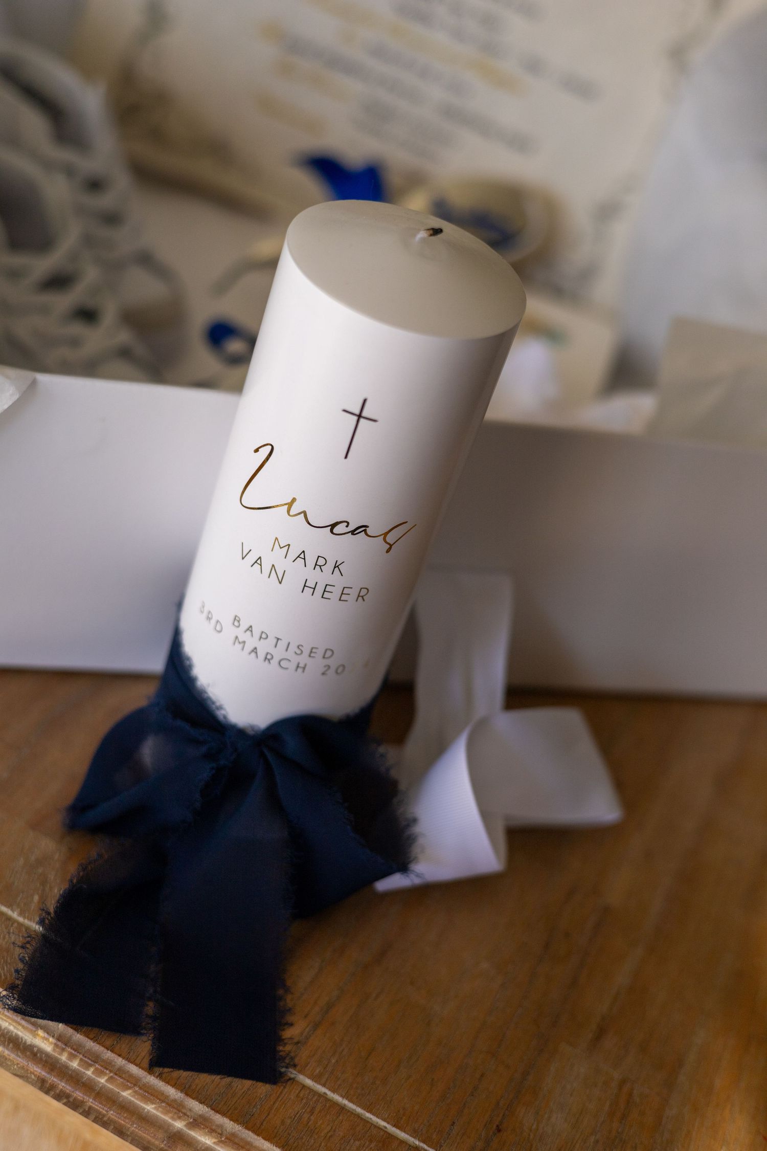White baptism or christening candle with cross symbol and navy blue bow ribbon on wooden surface.