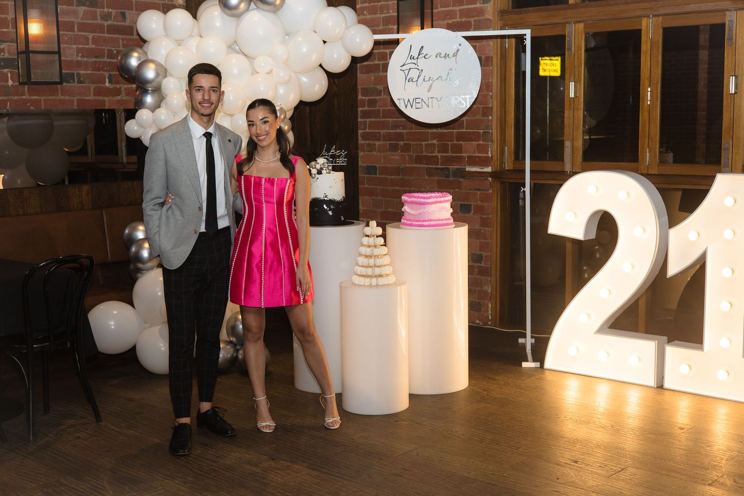 Two people pose by party decorations including white balloons, a cake display, and large illuminated '21' numbers.