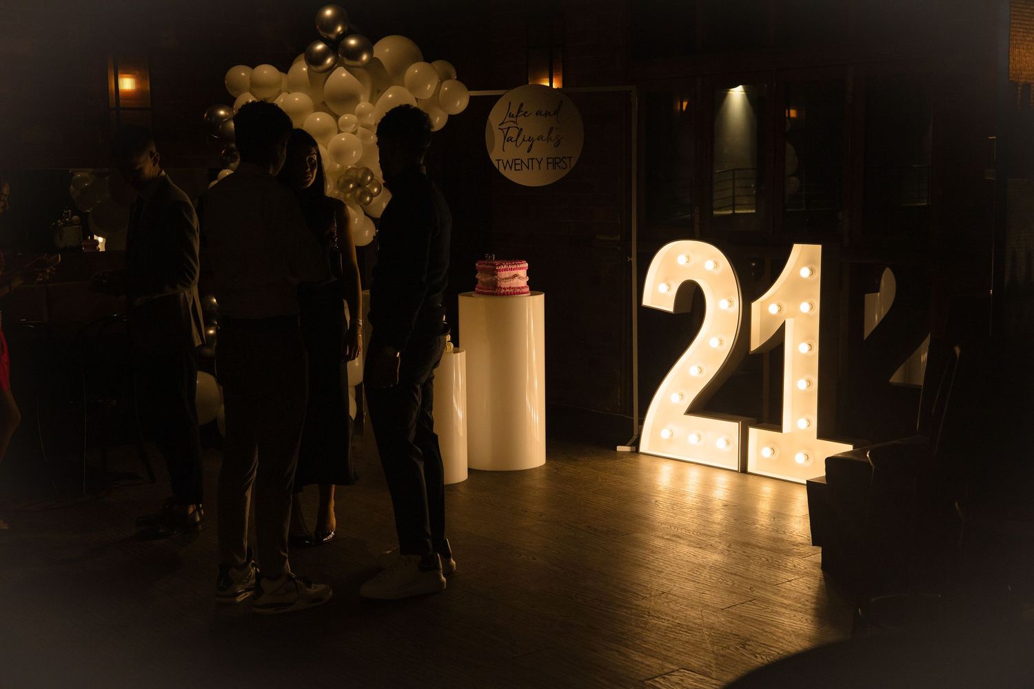 Illuminated number 21 display and silhouetted figures stand on a dark wooden floor at a celebration event.