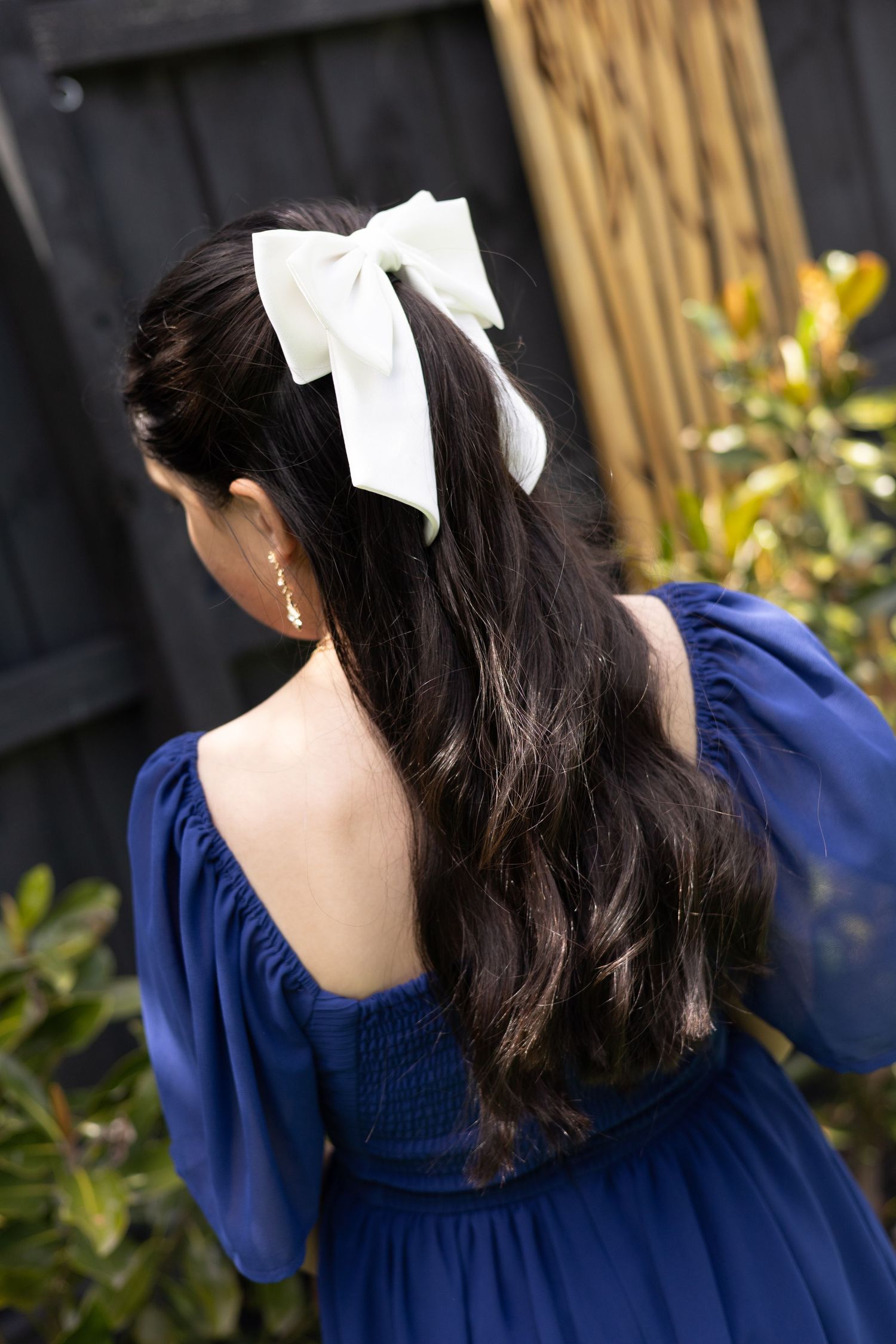Back view of dark wavy hair styled with a white bow accessory against a navy blue dress and garden backdrop.