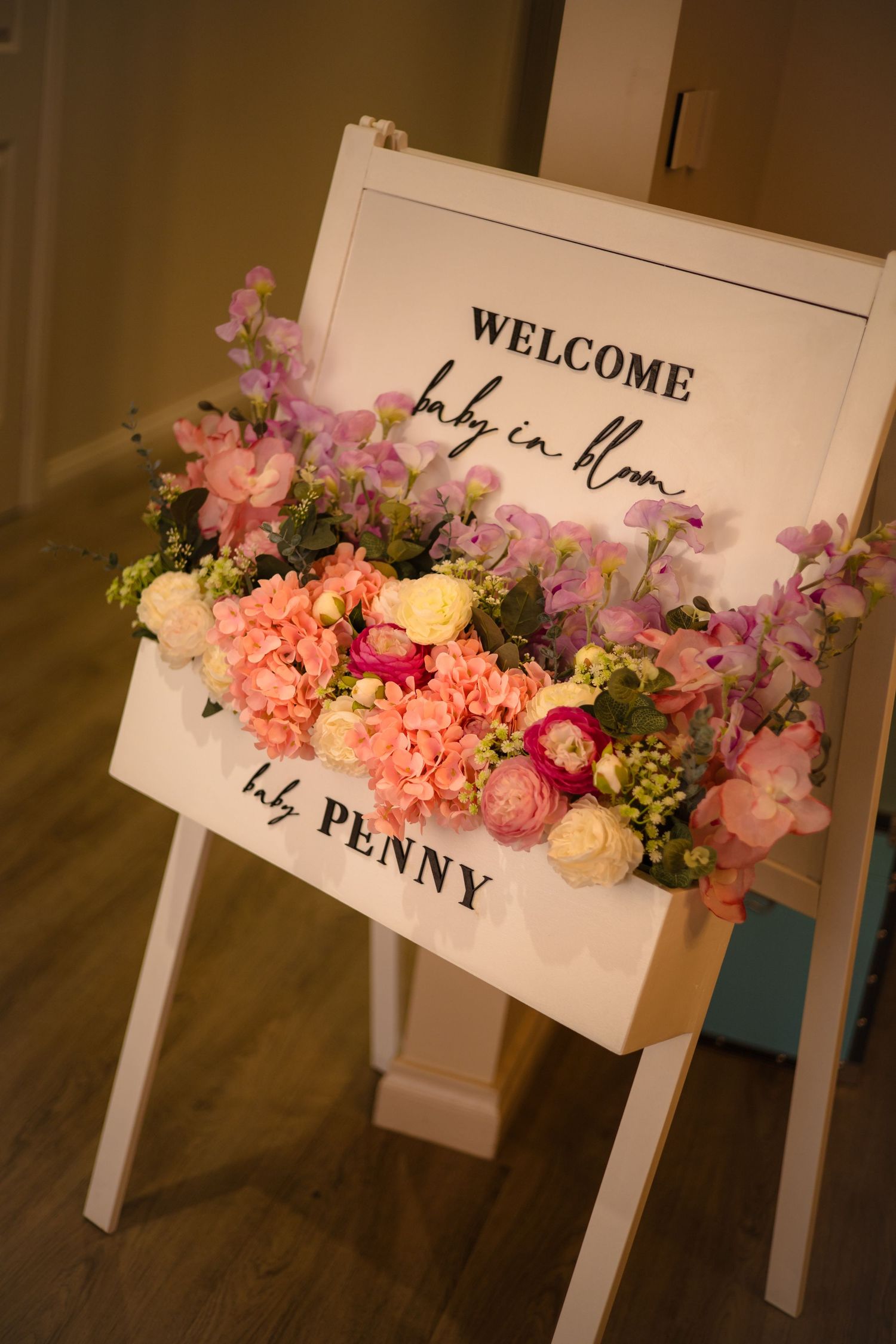 A white welcome sign decorated with pink, peach and yellow floral arrangements stands on an easel against a dark background.