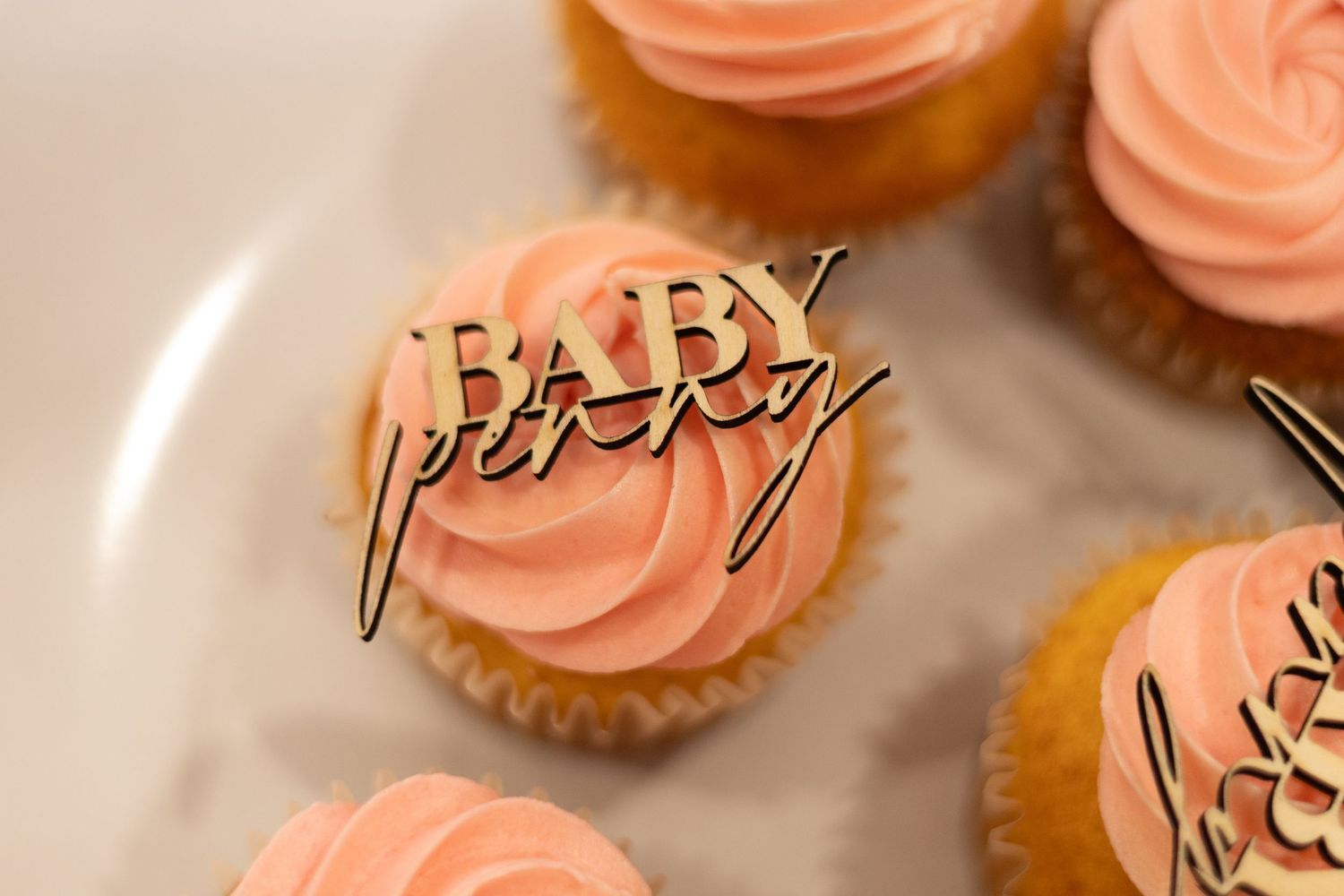 Elegant pink frosted cupcakes with golden 'Baby' text topper decoration on a white plate.