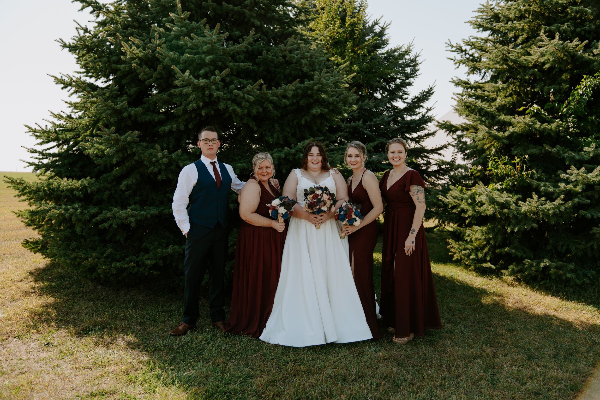 Wedding party poses on grassy hill with evergreen trees in the background.