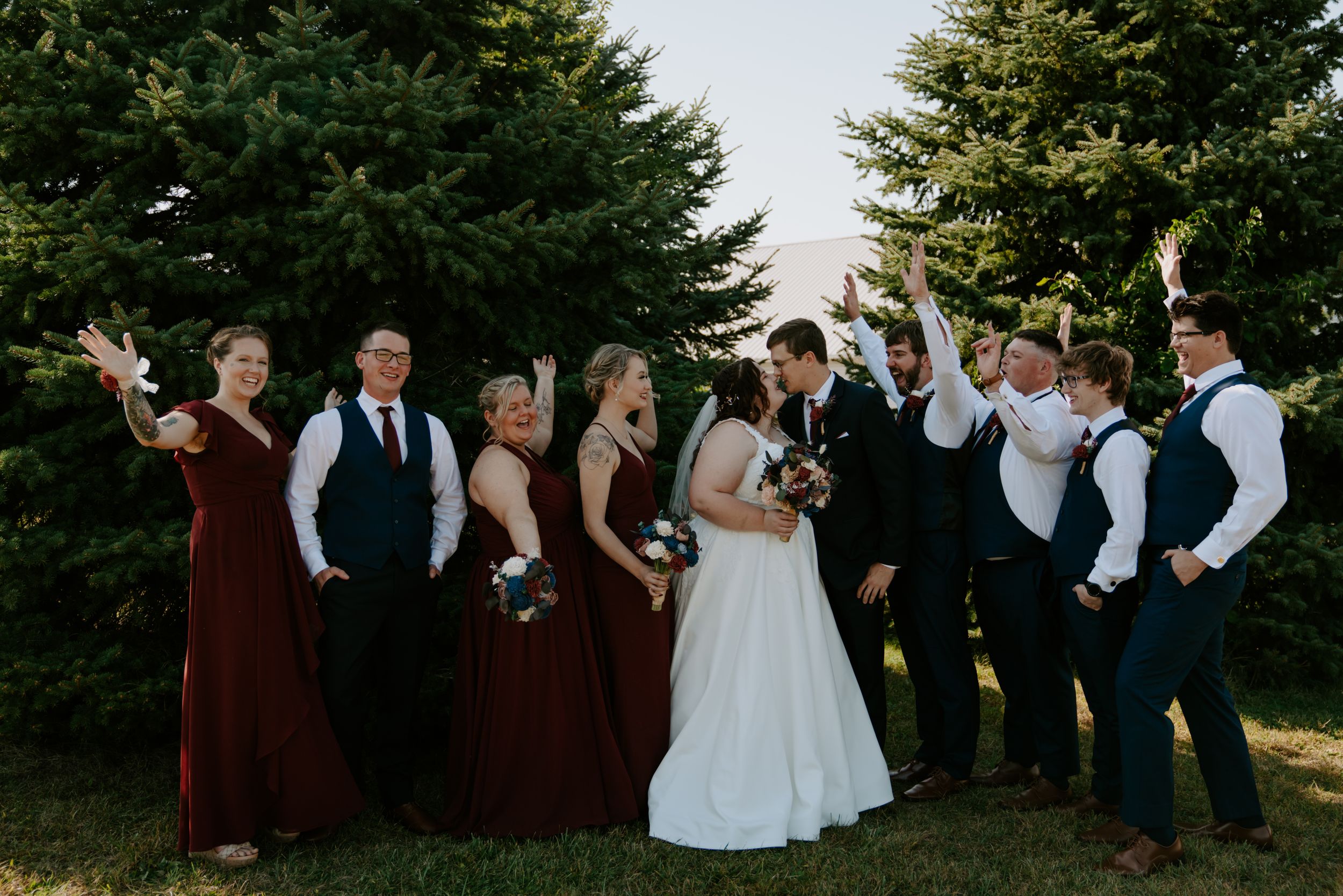 Formal wedding party in burgundy and black attire poses outdoors near pine trees.