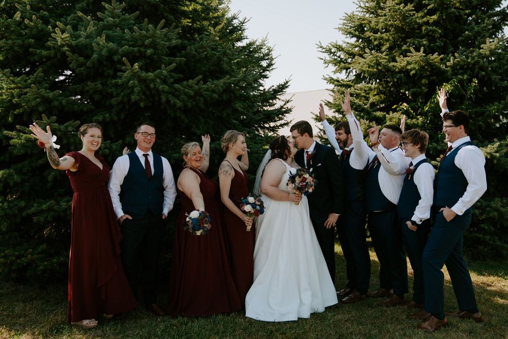 Formal wedding party in burgundy and black attire poses outdoors near pine trees.
