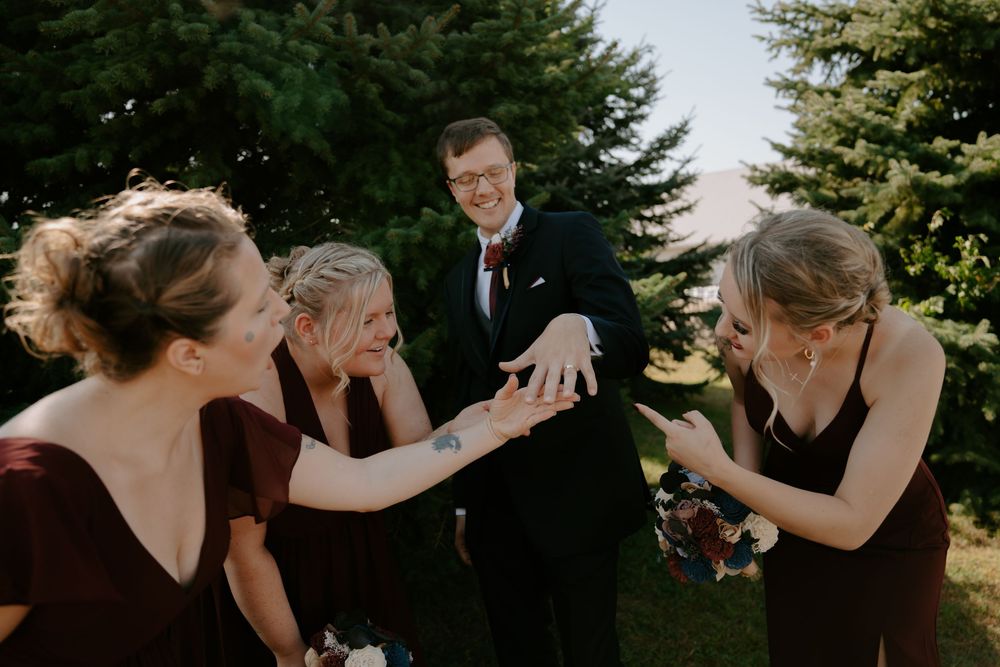 A joyful wedding party poses together outdoors in front of evergreen trees during a summer celebration.