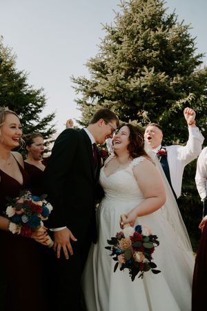Wedding party members celebrate with raised arms and laughter in front of pine trees.