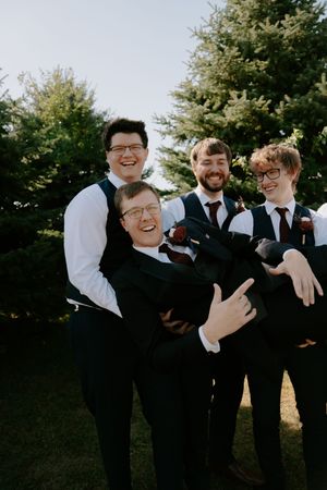 Groomsmen in black suits and white shirts pose energetically for wedding photos outdoors.