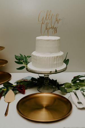 Simple white wedding cake displayed on gold platter with greenery accents.