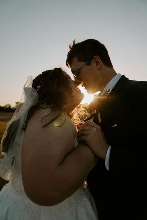 Romantic wedding couple shares intimate sunset kiss with sunburst behind them in outdoor setting.