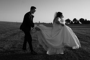 A couple in wedding attire poses in a field during sunset with flowing white dress fabric creating movement.