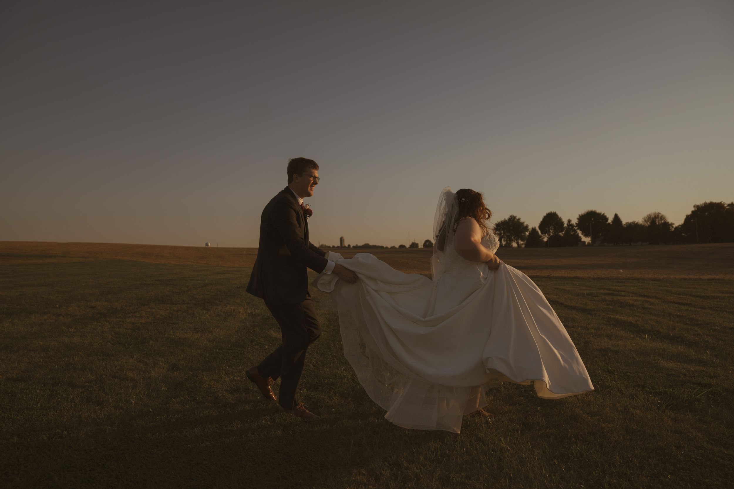 A couple in wedding attire dances in an open field at golden hour with dress fabric flowing in the wind.