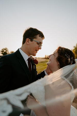 Bride's veil flows dramatically in wind as couple shares romantic moment at sunset.