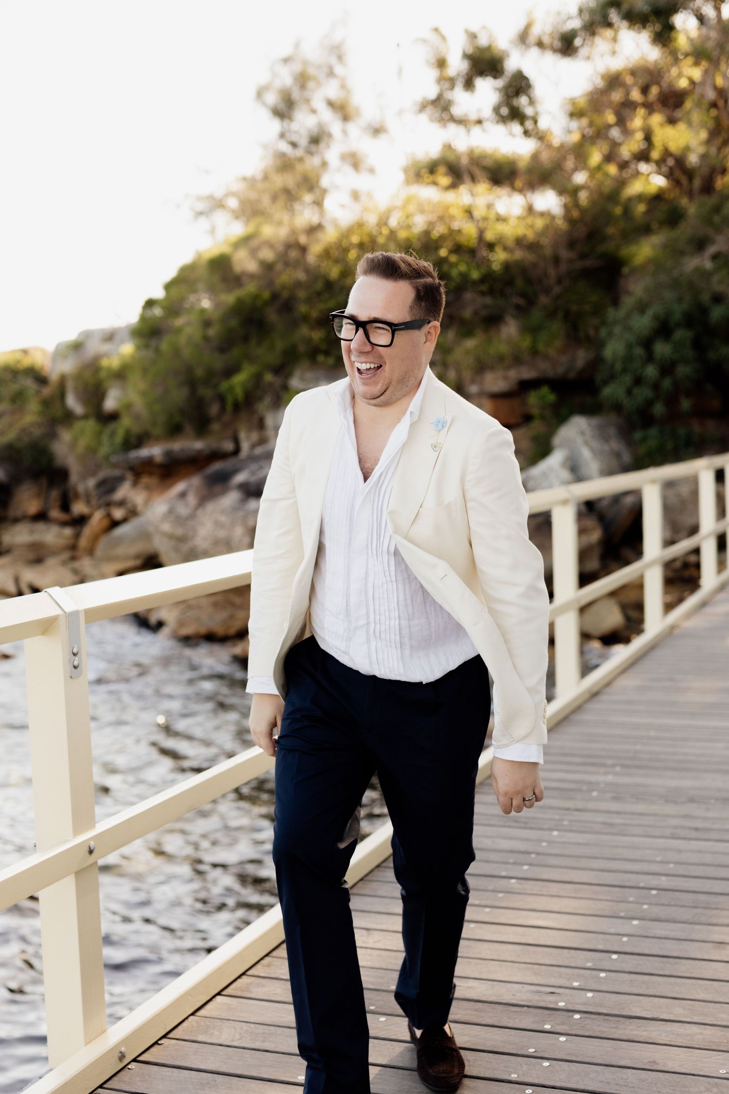 Smiling person in white blazer and black pants walking on wooden boardwalk by rocky shoreline at sunset.