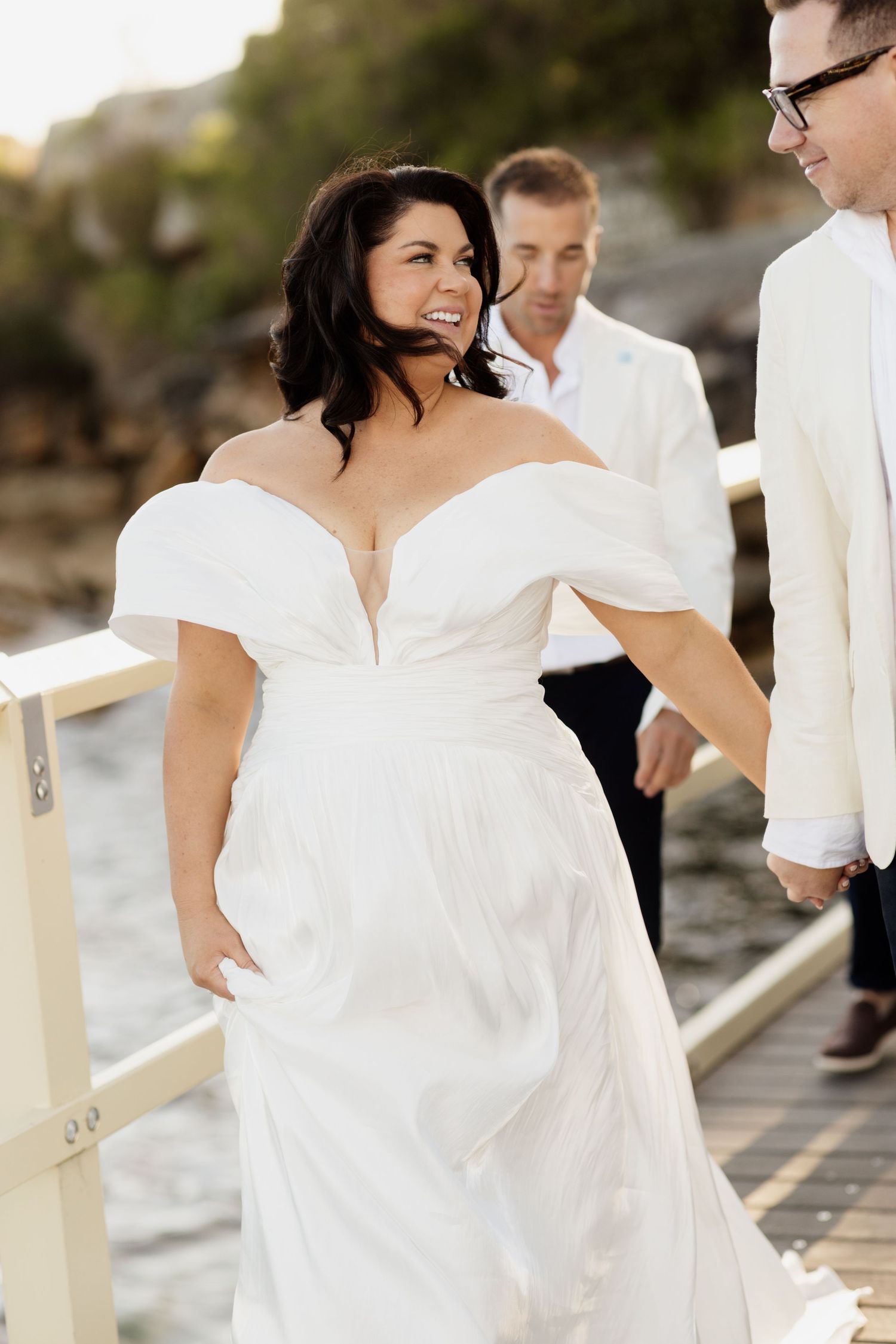 A bride in an elegant off-shoulder white dress walking along a seaside boardwalk on her wedding day.