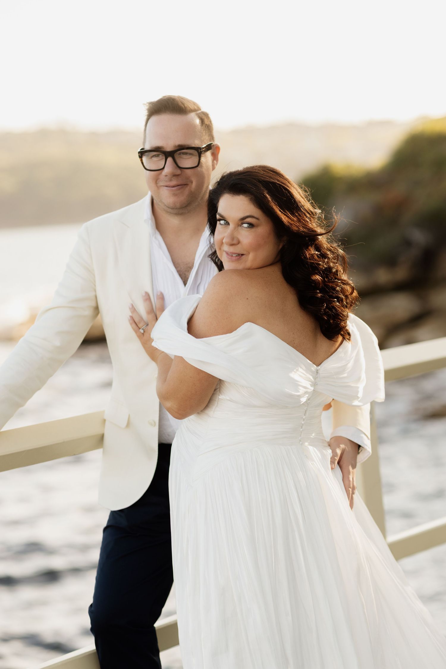 Romantic couple in wedding attire poses together on wooden pier during golden sunset by oceanside.