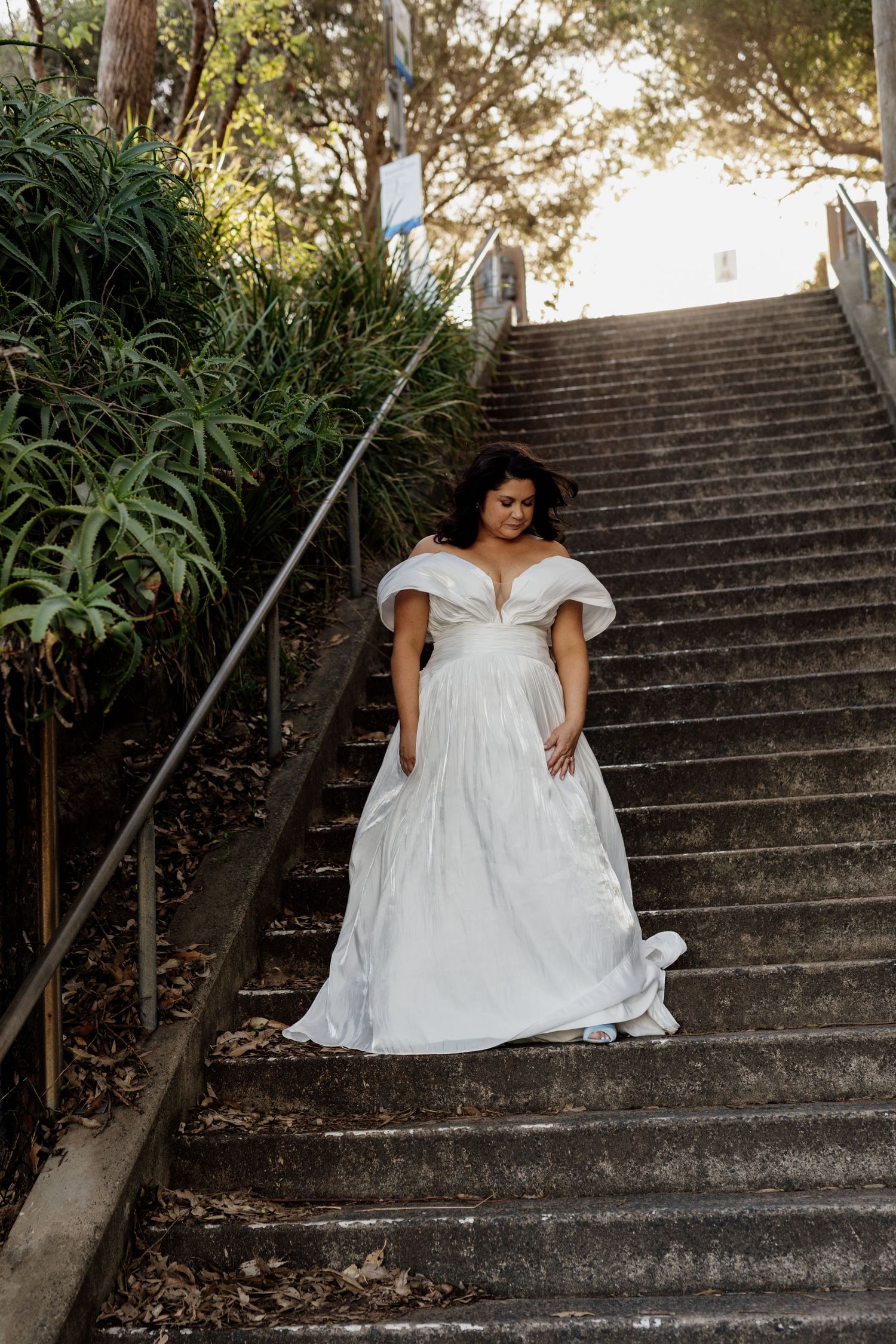 Figure in a flowing white dress sits gracefully on concrete steps surrounded by lush greenery at sunset.