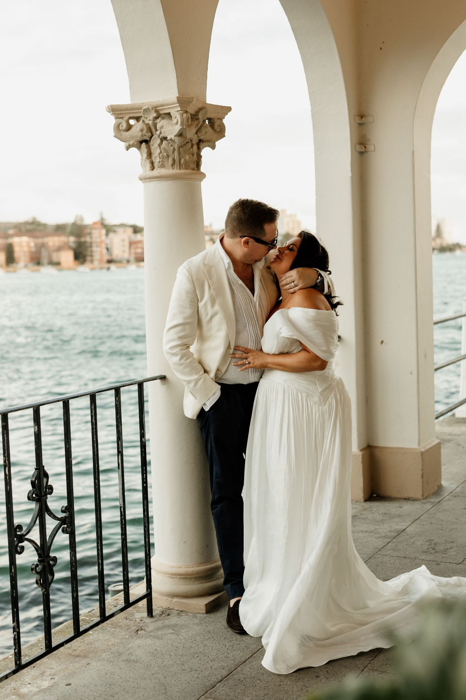 A romantic couple shares an intimate moment on a waterfront balcony with white columns and ornate architecture in Venice.