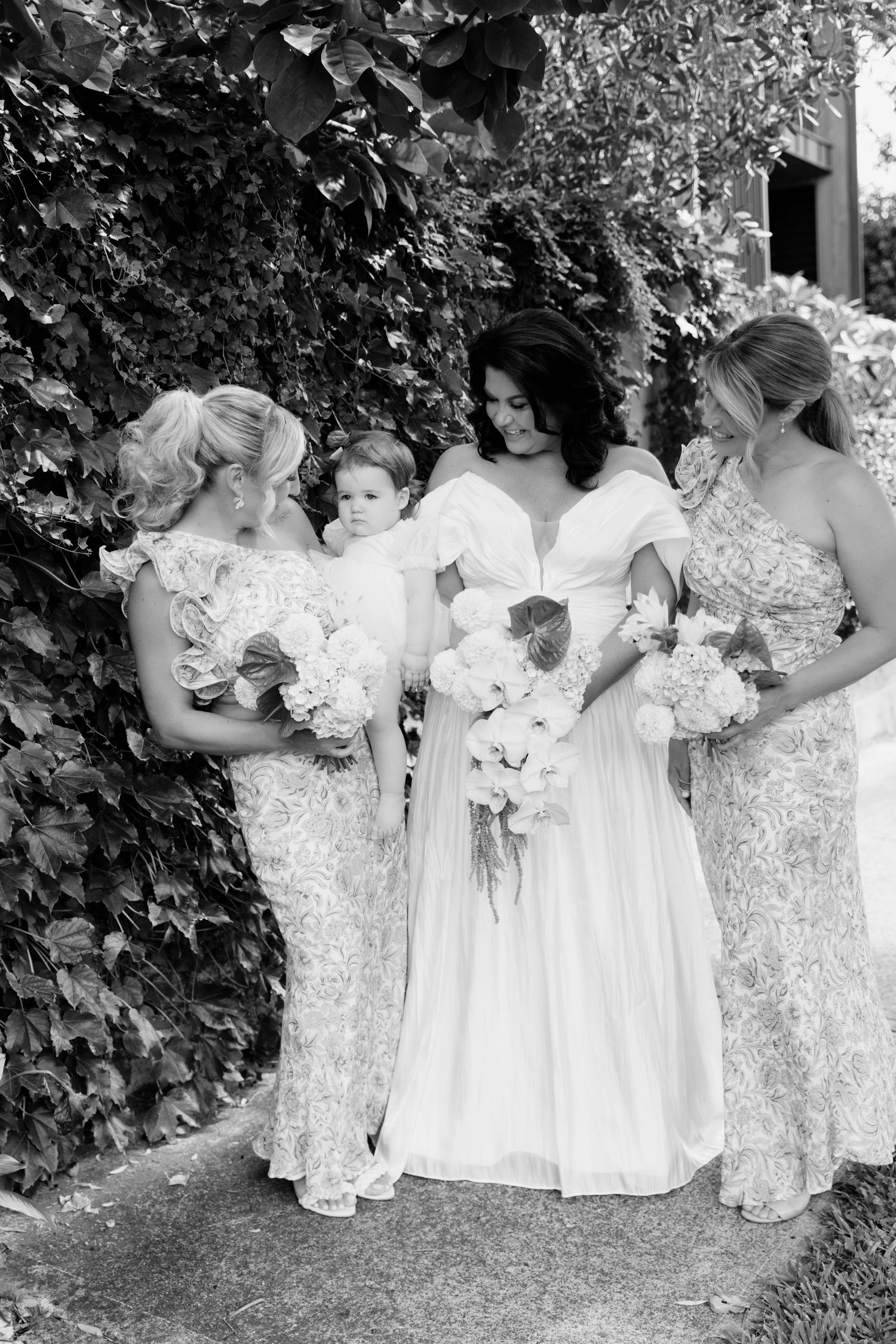 A bride in white and bridesmaids in lace dresses gather together holding bouquets in a garden setting.