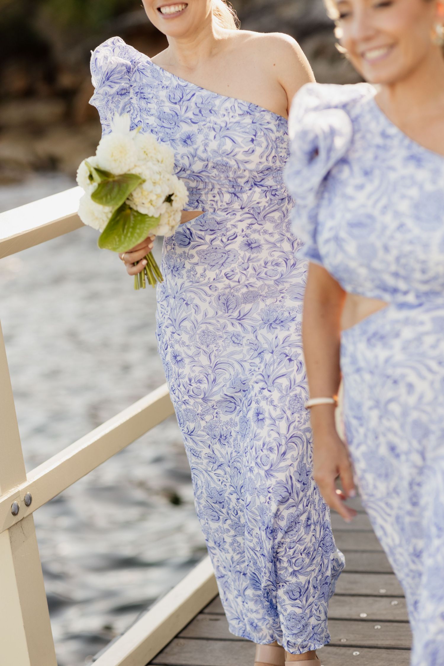 Bridesmaids wearing elegant blue and white floral one-shoulder dresses hold white bouquets on a beach boardwalk.