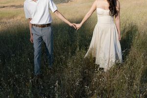 A couple holds hands while standing in a sunlit meadow filled with tall grass.