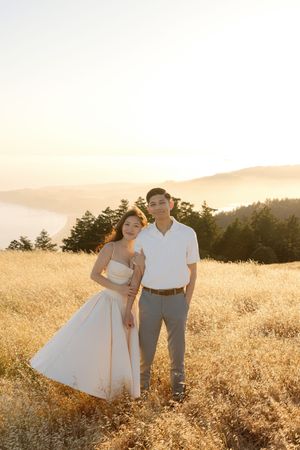 A couple stands in a golden field at sunset with mountains and trees in the background.