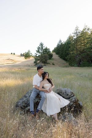 A couple sits intimately on a large rock surrounded by golden grass and evergreen trees in a scenic hillside setting.