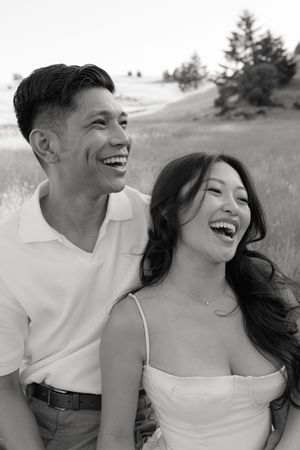 A joyful couple shares genuine laughter together in a grassy field with evergreen trees in the background.