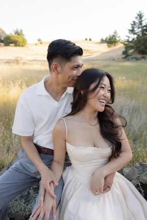 A romantic couple sits together in a golden grass field during sunset wearing elegant white attire.
