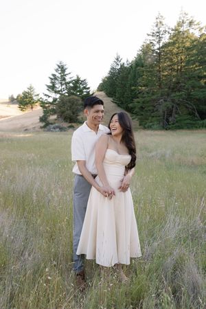 A couple shares a joyful moment together in a scenic field with evergreen trees in the background.