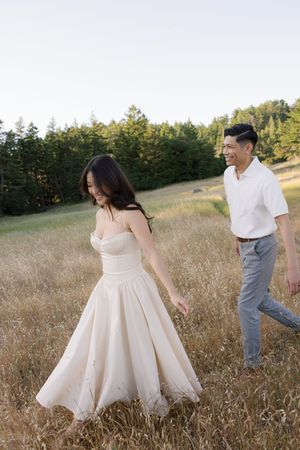 A couple in light-colored clothing walks through a sun-drenched field with pine trees in the background.