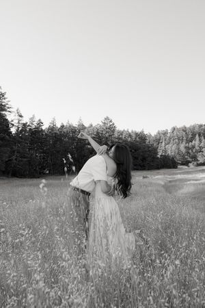 Black and white photograph of a person in a flowy dress dancing in a meadow with trees in background at sunset.