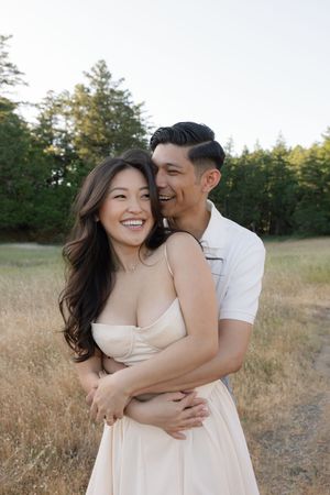 A romantic couple embracing in a golden field at sunset with pine trees in the background.
