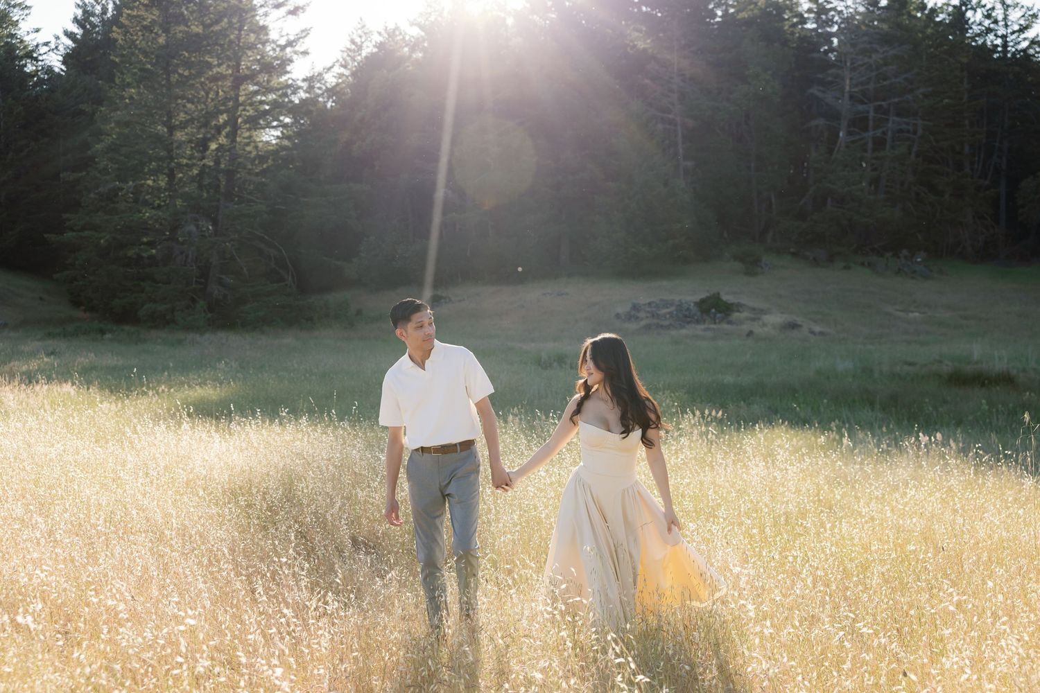Two people stand together in a sunlit golden meadow surrounded by evergreen trees.