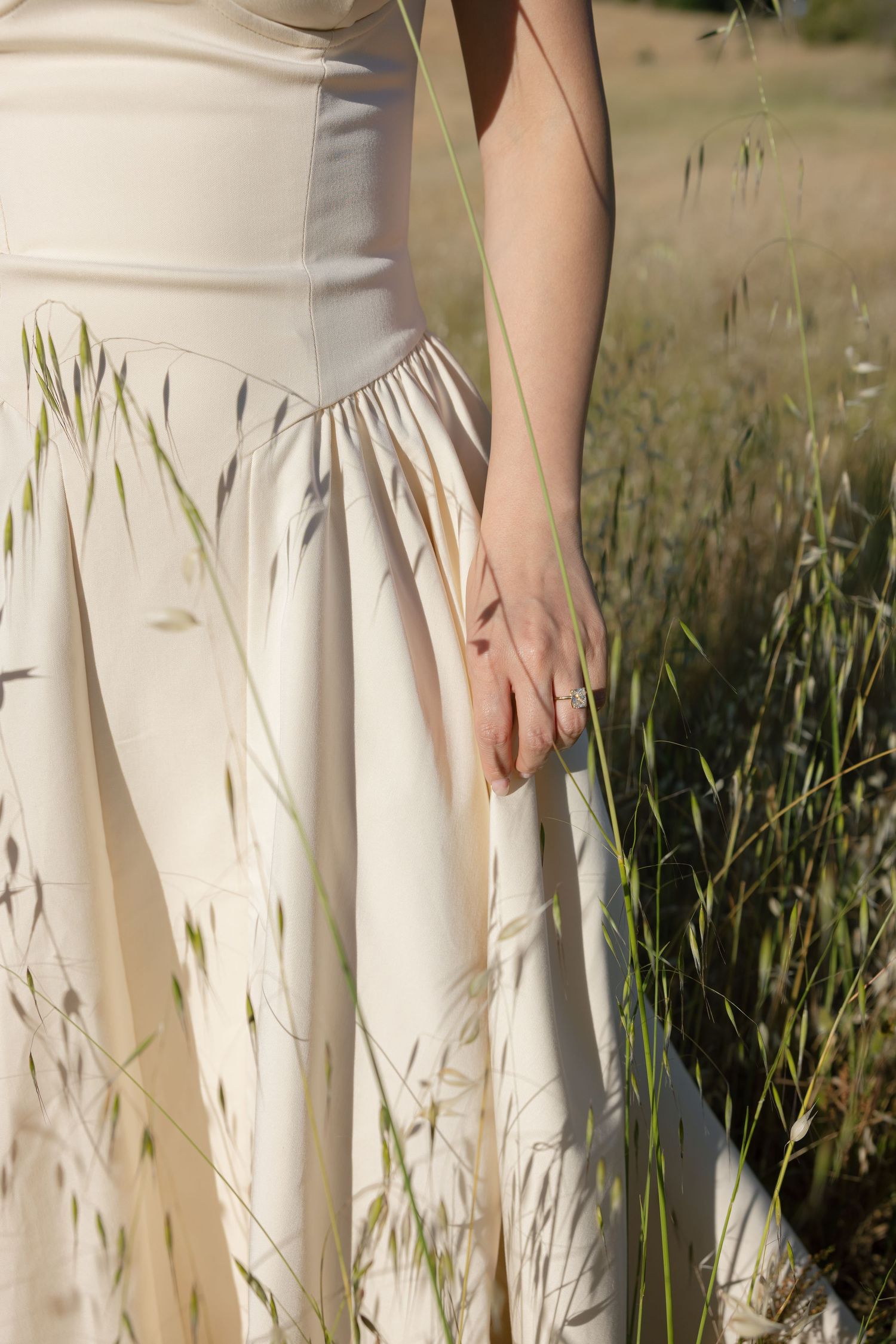 Close-up detail of a cream-colored dress flowing in tall grass against natural sunlight.