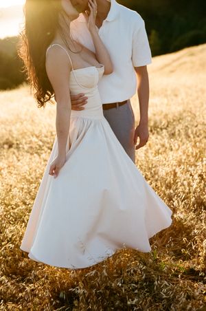 Couple in a romantic pose wearing white dress and casual attire standing in a golden sunlit field at sunset.
