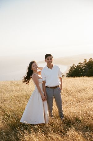 A couple in white clothing poses together in a golden sunlit field during sunset with trees in the background.