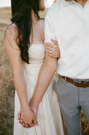 A romantic couple holding hands outdoors, with focus on white dress and ring detail.