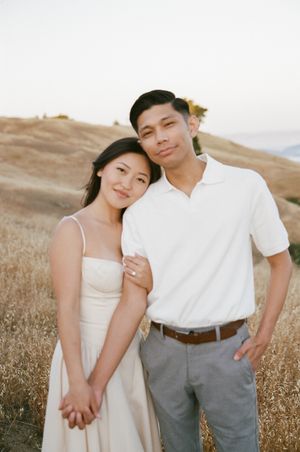 A couple in romantic casual attire poses together in a sun-dried grass field during golden hour.