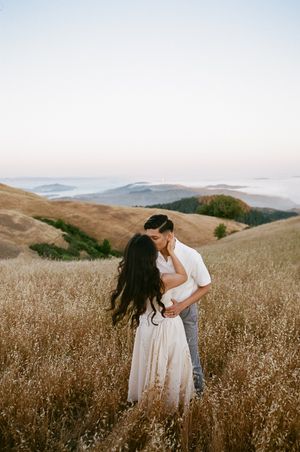 Couple sharing an intimate moment in a golden grass field with misty mountains in background.
