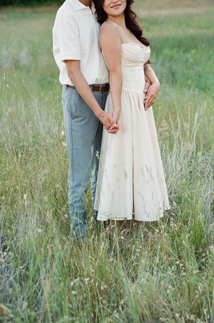 A couple stands together in a tall grass meadow wearing a white dress and gray slacks on a sunny day.