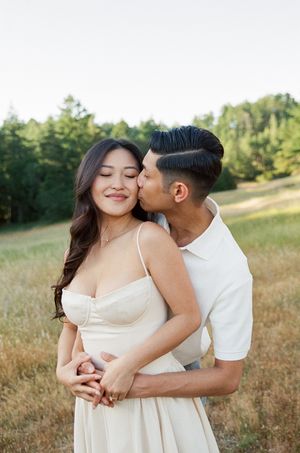 A romantic couple embraces in a golden field at sunset, she wearing a white sundress as they share an intimate moment.