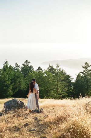 A couple embraces in a golden field at sunset with evergreen trees and a large boulder in the background.