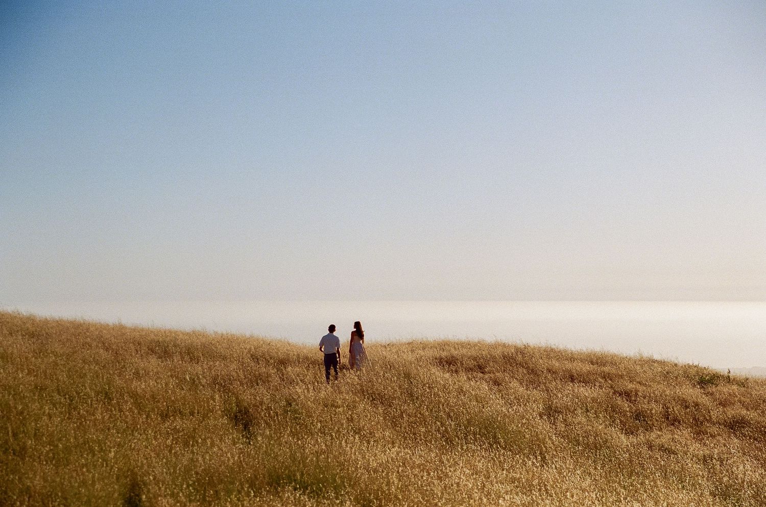 Two silhouetted figures walk along a grassy hilltop against a hazy sky during golden hour.