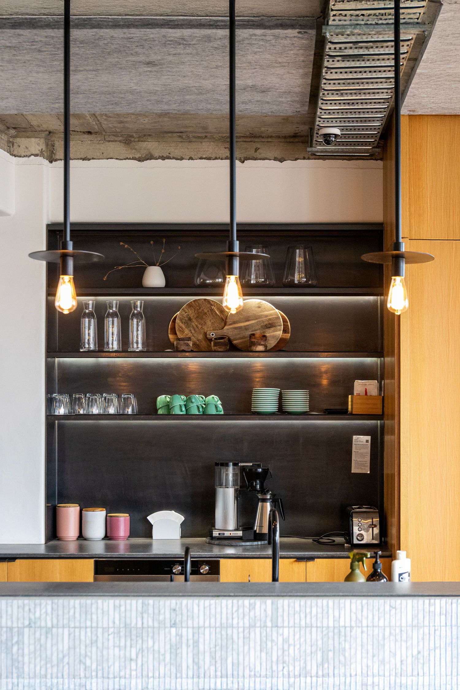 Warm-toned café interior with timber shelving, hanging bulbs, and ceramic mugs neatly arranged above the coffee counter.