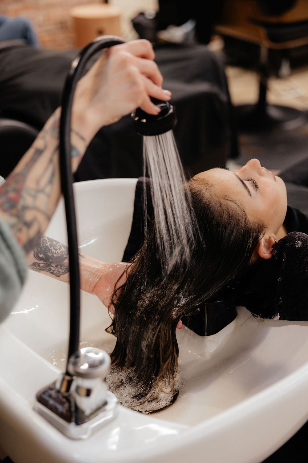 Client with dark hair gets a relaxing shampoo at a salon sink while resting with hair being rinsed by gloved hands.