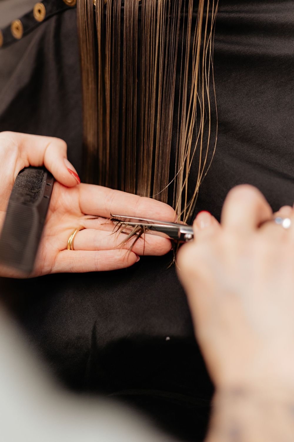 Close-up view of hands trimming long brown hair with professional scissors against dark background.