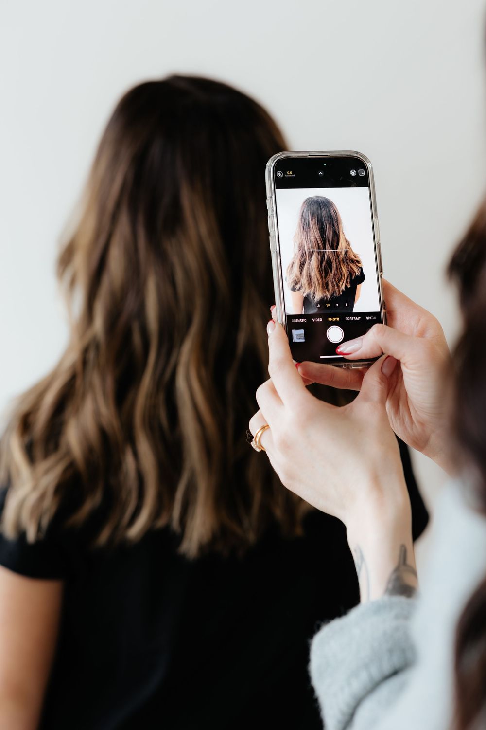 A person holding a phone to take a photo of their dark brown shoulder-length hair against a white wall.